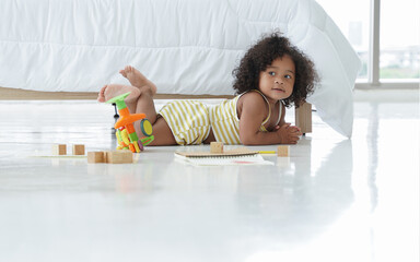 Portrait of happy African little kid girl with curly hair smiling and lying on the floor playing toys wooden blocks, drawing book at bedroom at home - Powered by Adobe