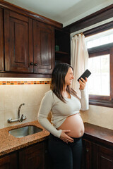 Vertical shot of a pregnant Hispanic woman recording audio on her cell phone while holding her belly and looking out the kitchen window.