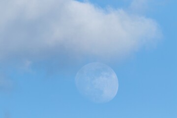 A portrait of a blue sky with a dark grey cloud in it and below the cloud the moon is faintly present.