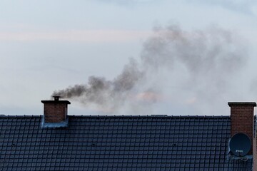 A roof with two chimneys on it. In one of the chimneys there is a pipe mounted on it were a lot of black polluting smoke is coming out or rising up from, connected to the fireplace of the house.