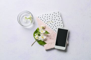 Mobile phone, notebook, jasmine flowers and glass of water on light background