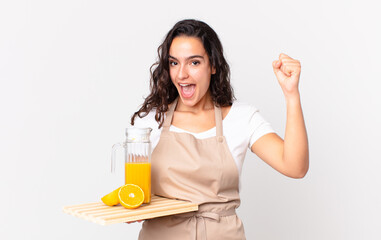 hispanic pretty chef woman feeling shocked,laughing and celebrating success and holding an orange...