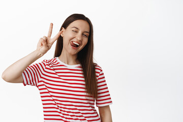 Fototapeta premium Happy young woman wink, show tongue silly and peace v-sign, positive summer mood, wearing striped red t-shirt, standing against white background