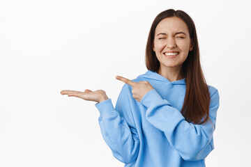 Happy cute girl in hoodie, laughing and smiling, pointing finger at empty open palm, showing something in her hand, standing against white background