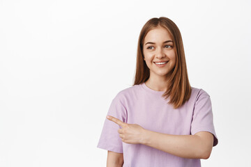 Fototapeta premium Portrait of cute blond girl, student in t-shirt pointing, smiling and looking left at promo text, showing banner, event or store advertisement on empty space, white background