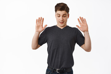 Fototapeta premium Surprised young man drop something, raising hands up and looks down startled, staring below, standing against white background in casual clothes