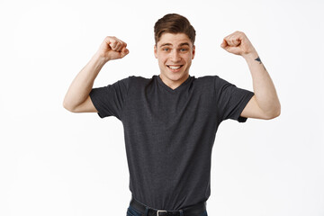 Fototapeta premium Excited young man winning, chanting and celebrating victory, raising hands up in hooray triumph gesture, achieve goal and fist pump, standing happy against white background