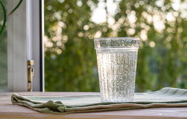 A glass glass with mineral water with gas bubbles stands on a wooden window sill with the window open to the street. Topic thirst quenching, a healthy natural drink