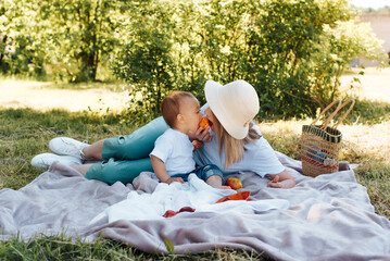 Family portrait of a beautiful young mother in hat and little son, outdoors. Mom and child have fun eating one fruit together, lying on blanket on the lawn summertime. Family picnic in nature concept. - obrazy, fototapety, plakaty