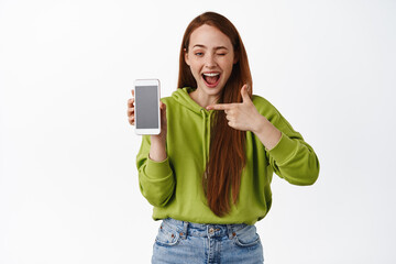 Cheeky ginger girl showing app, pointing at smartphone empty screen and winking at camera happy,...
