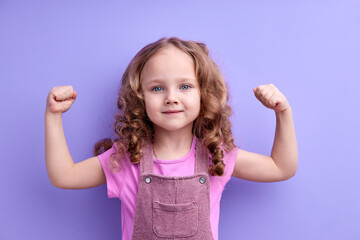 Happy caucasian little child girl showing arm muscles looking at camera isolated on purple background with copy space. Strong kid in casual outfit with good health concept