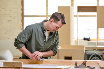 Male carpenter working with equipment on wooden table in carpentry shop, polishing the wooden material, making house furniture, using tools. woodworking, carpentry concept