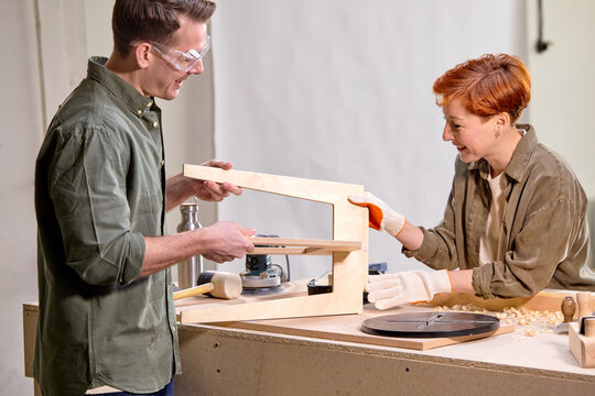 Team work of two friendly joiners working in workshop together, carpentry concept. young man and woman discuss what to do with new stool chair, in workshop. side view portrait. copy space