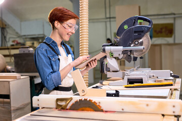 Beautiful caucasian female carpenter with red hair using digital table at work standing next to...