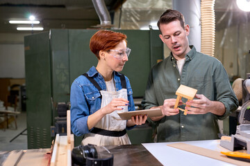 Two carpenters working with equipment on wooden table in carpentry shopm discuss design project of future stool using digital tablet. Busy woman and man tinkering in workshop, having talk