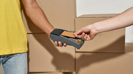 Close up shot of hands of delivery man holding terminal for a customer to pay for parcels received from courier with credit card. Cardboard boxes in the background