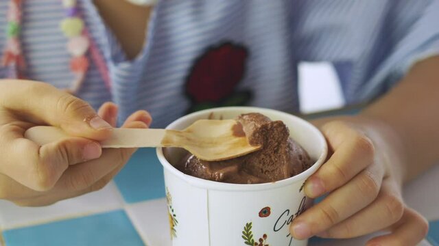 Close up 4K of little child is eating delicious and tasty dessert (homemade chocolate ice cream) happily which looks like soft gelato in single use cup bowl and wooden spoon in cafe.
