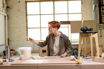 Young Caucasian Carpenter Handywoman Painting Wooden Plank In Workshop, Portrait Of Redhead Female In Casual Shirt Engaged in Carpentry Work, In Workshop Alone, Side View. Handicraft Concept