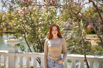 Sensual young redhead woman standing in casual wear posing next to blossoming tree in spring garden. Natural Beauty and nature consept. Caucasian model looking at camera. Copy space