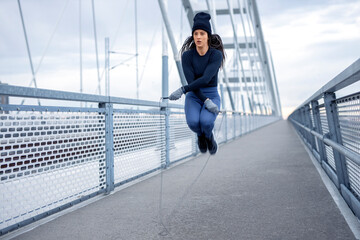 Young fitness woman in winter blue sportswear doing exercise with jump rope on bridge during cold day