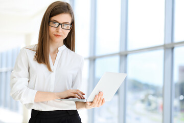 Beautiful female specialist with laptop computer standing in modern office and smiling charmingly. Working on design, data analysis, plan strategy. Business people concept