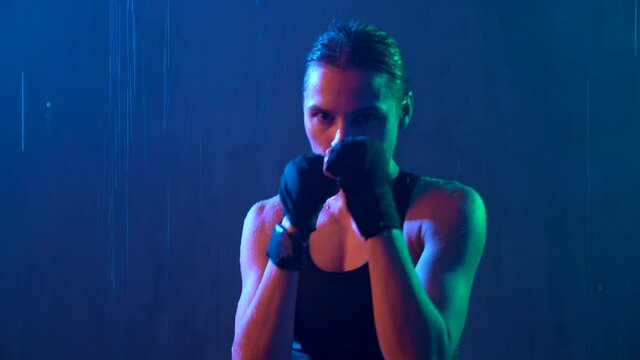 Side view of muscular girl with sports bandages on hands doing hit box workout. Close up motion of young sportswoman training in rainy neutral dark atmosphere, looking at camera, trends 2020.