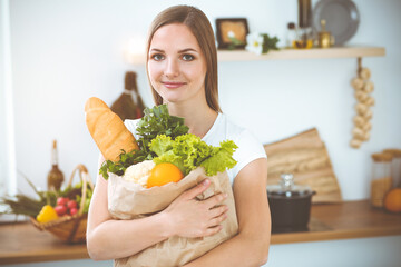 An attractive young woman holding the paper bag full of vegetables while standing and smiling in sunny kitchen. Cooking concept