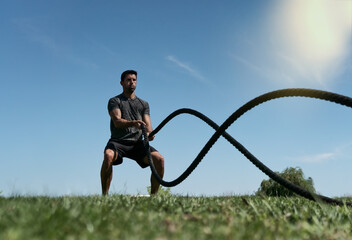 Male athlete on the outdoor in the park, exercises for endurance, using a thick rope, for cross...