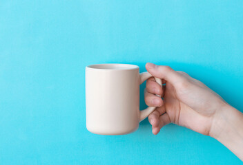 Human hand holding big ceramic beige mug above light blue background with copy space. Mug mock up for your design