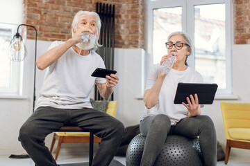 Retired man and woman refreshing with water after domestic workout. Senior couple in sport clothes using smartphone and tablet during relaxation.