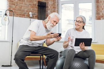 Smiling mature man and woman taking break during morning exercises and using modern tablet with smartphone. Concept of people, recreation and gadgets.