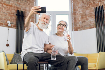 Cheerful mature couple in activewear taking selfie on smartphone while resing with bottle of water after training. Concept of retirement, sport activity and technology.