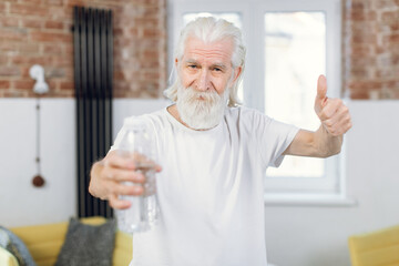 Bearded senior man showing thumb up while holding bottle of fresh water. Male in activewear renewing water balance after workout at home.