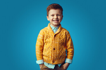 Kid smiling while holding hands in pockets, isolated over blue background