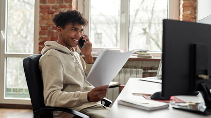 Successful young male trader holding papers, smiling while talking on the phone with a client, sitting at desk, trading from home