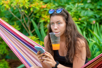 gorgeous girl sitting on the hammock relaxing, drinking orange juice and using smartphone