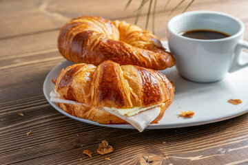 Two French croissants with ham and cheese and a cup of black coffee on the white plate. Wooden table, morning country breakfast.