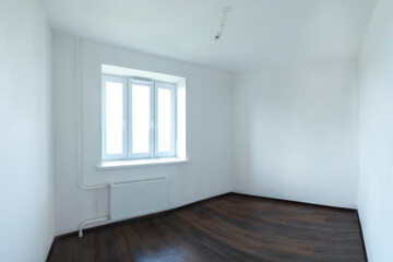 Panoramic photo of an empty clean room with white walls, a white radiator and a dark laminate floor. A light bulb on the ceiling. A closed window. Three corners of the room are visible.