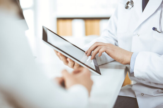 Unknown male doctor and patient woman discussing current health examination while sitting in clinic and using tablet computer. Perfect medical service in hospital. Medicine and healthcare concept