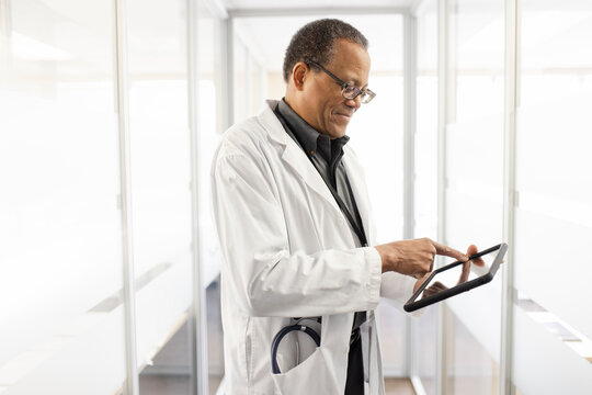 Male Doctor In Lab Coat Using Digital Tablet In Clinic Corridor