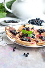 Breakfast with tea, pancakes, fresh blueberries and mint on grey background. Fried pancakes on a white plate with fork and knife.