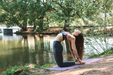 Young attractive woman practicing yoga,Camel pose, in the park on the lake shore.Sports healthy lifestyle.Outdoor training.Slow living concept.Copy space.