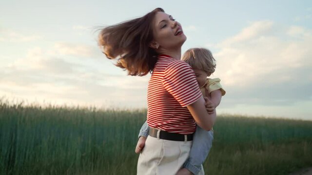 Happy family - mom and baby son spinning around, smiling, having fun on nature field background. Summer season. European young mother and 2 years old toddler child.