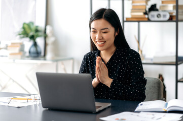 Asian happy young woman, sit at workplace, rejoicing at success, victory, good result, palms folded together in front of her. Chinese or Japanese woman, greets interlocutor during an online video call