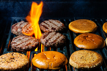 Grilling round patties of ground beef or pork and burger buns on a grill. Tongues of flame are...