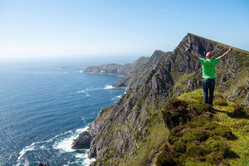 Bald male tourist in green shirt hands up in the air Beautiful scenery of Achill island, county Mayo, Ireland. Warm sunny day, blue sky and water. Travel concept.