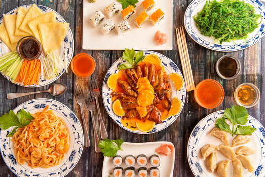Top view image of a plate of food cooked in a Chinese restaurant with udon noodles, orange duck, Pekin duck, salmon makis, chopsticks, gyozas, wakame seaweed salad and all kinds of sauces