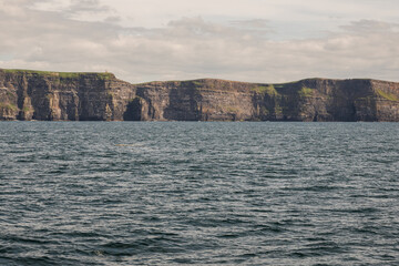 Fototapeta na wymiar View on Cliff of Moher from a board of a ferry or cruise ship. West coast of Ireland. Popular tourist destination. National landmark. Cloudy sky. Calm surface of Atlantic ocean.