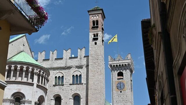 Trento, Trentino, Italy, June 2021. On a beautiful summer day nice pan footage of the San Vigilio Cathedral rear three quarters. Next to it the bell tower surmounted by a yellow flag.