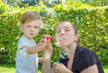 Cute little boy and his mother having fun blowing soap bubbles in the park. Funny outdoor activity for children in a beautiful summer sunny day